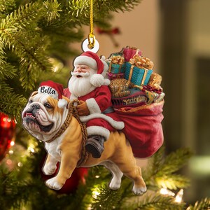 Pu&ograve; includere: Un ornamento natalizio con Babbo Natale che cavalca un bulldog. Babbo Natale indossa un abito e un cappello rossi, con barba bianca e un sacco di regali. Il bulldog &egrave; marrone e bianco, con un cappello da Babbo Natale con il nome "Bella".
