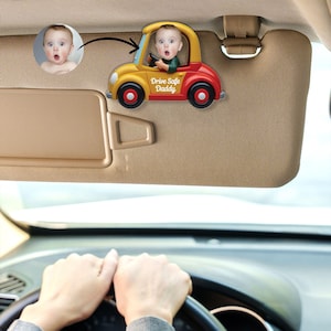 May include: A yellow and red toy car with a baby's face in the driver's seat. The car has the words "Drive Safe Daddy" on the side. The car is attached to a car sun visor.