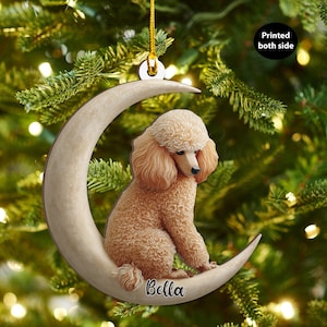 May include: A beige poodle Christmas tree ornament sitting on a crescent moon. The ornament has a gold string for hanging. The text "Bella" is written on the moon. The background is a blurred Christmas tree.