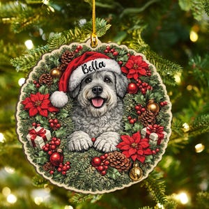 May include: A round wooden Christmas ornament featuring a gray dog wearing a red Santa hat with the name Bella printed on the brim. The dog is surrounded by a wreath of green pine needles, red poinsettias, pinecones, red berries, and gift boxes.