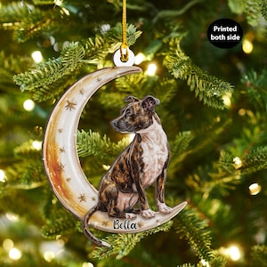 May include: A dog-themed Christmas ornament featuring a brindle and white dog sitting on a crescent moon with star accents. The ornament has the name "Bella" and the text "Printed both side". The ornament is hanging on a Christmas tree.