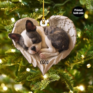 Può includere: Un ornamento natalizio a forma di cuore con un cucciolo di Boston Terrier addormentato in ali d'angelo. Il cucciolo ha segni neri, bianchi e marroni. Il nome "COCO" è stampato sull'ornamento. Il testo "Printed both side" è anche visibile.