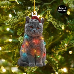 May include: A gray cat Christmas ornament wearing a Santa hat with the name "Bella" and wrapped in colorful Christmas lights. The ornament is hanging from a gold string against a backdrop of a Christmas tree.