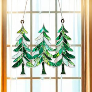 May include: Three stained glass pine trees hanging in a window. The trees are green and white with a silver chain.