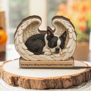 Op de afbeelding: Een houten herdenkingsornament met een slapende zwart-witte Boston Terrier hond genesteld in een paar uitgesneden engelvleugels. De naam Bella is in de vleugel gegraveerd en de basis toont de tekst, You may have left my arms, but you'll never leave my heart.