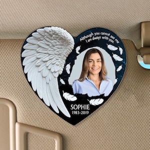 Puede incluir: Un amuleto de coche conmemorativo en forma de corazón negro con un ala de ángel blanca, una foto de una mujer y el texto "Aunque no puedas verme, siempre estoy contigo" y "SOPHIE 1983-2019".