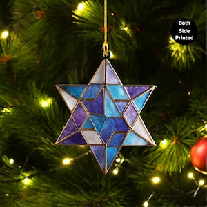 Puede incluir: Un adorno navideño en forma de estrella con un diseño geométrico. El adorno presenta un mosaico de piezas de vidrio azules, moradas y blancas, enmarcado por un metal dorado. Cuelga de un hilo dorado sobre un fondo de árbol de Navidad.