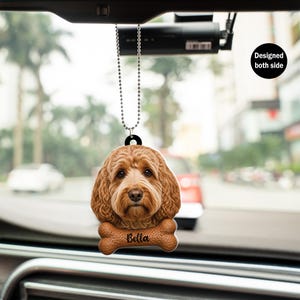 May include: A brown dog car charm with the name "Bella" on a bone-shaped accent. The charm is designed to hang from a chain and is visible from both sides. The background shows a blurred street scene.