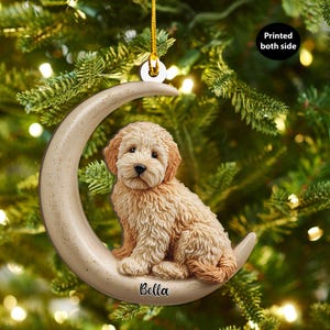 Può includere: Un ornamento a forma di mezzaluna beige con un cane Goldendoodle marrone chiaro. Il cane ha una pelliccia soffice e un'espressione dolce. Il nome "Bella" è scritto sulla luna. L'ornamento è appeso a un filo d'oro e ha il testo "Printed both side".