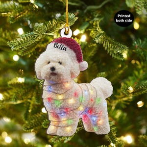 Puede incluir: Adorno navideño de perro Bichon Frise blanco con gorro de Papá Noel con el nombre "Bella" y una cadena de luces de colores. El adorno cuelga de un hilo dorado sobre un fondo de árbol de Navidad.
