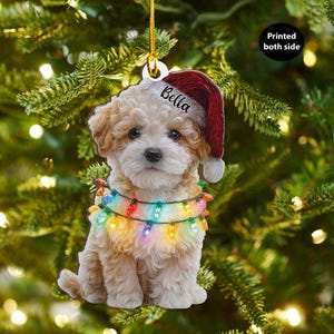 Può includere: Un ornamento natalizio raffigurante un cucciolo marrone chiaro e soffice con un cappello da Babbo Natale con il nome "Bella" e una ghirlanda di luci natalizie colorate. L'ornamento è appeso davanti a uno sfondo sfocato di albero di Natale. Il testo "Printed both side" è visibile.