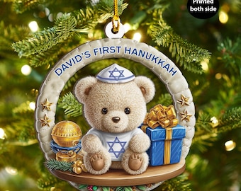 Può includere: Ornamento natalizio rotondo con un orsetto che indossa una kippah e una maglietta con una stella di David. L'ornamento riporta la scritta "David's First Hanukkah" e include un pacco regalo blu e un globo dorato. È appeso a un filo d'oro.