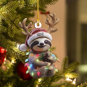 Puede incluir: Adorno navideño con un perezoso con un gorro de Papá Noel con el nombre "STEVE" y cuernos de reno. El perezoso está envuelto en luces navideñas de colores y sostiene una piña. El adorno cuelga de una cuerda dorada.