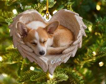 May include: A heart-shaped Christmas ornament featuring a sleeping corgi dog nestled within angel wings. The ornament is tan and gold, with the dog's fur in shades of brown and white. The name "Bella" is inscribed on the ornament.