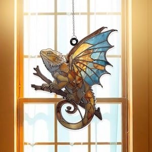 May include: A stained glass-style suncatcher depicting a lizard with wings, perched on a branch. The lizard has a tan body, blue and yellow wings, and is suspended by a chain. The background is a window with natural light.