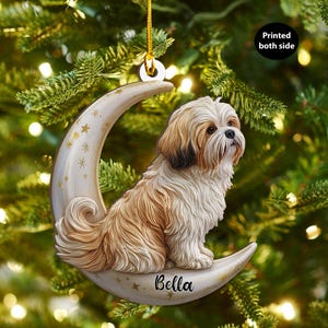 May include: A decorative Christmas ornament featuring a Shih Tzu dog perched on a crescent moon. The ornament is cream-colored with gold stars and the name "Bella" on the moon. The dog has brown and white fur.