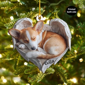 Könnte beinhalten: Herzförmiger Weihnachtsschmuck mit einem schlafenden Corgi-Hund mit Engelsflügeln. Der Hund ist braun und weiß, eingebettet in ein Federbett. Der Name "Bella" steht unten. Der Schmuck ist beidseitig bedruckt.