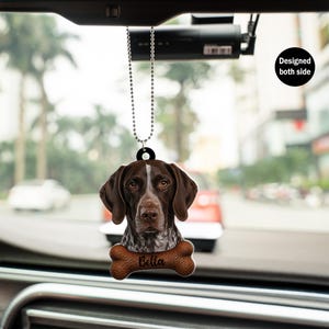 May include: A car hanging ornament featuring a brown and white dog with the name "Bella" on a bone-shaped tag. The ornament is designed to be visible from both sides and hangs from a beaded chain.