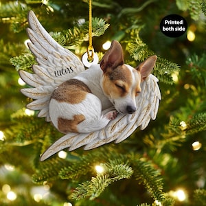 May include: A Christmas tree ornament depicting a sleeping dog within angel wings. The dog has brown and white fur, and the wings are off-white with gold accents. The name "LUCAS" is printed on the wing. The text "Printed both side" is also visible.