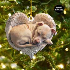 Puede incluir: Adorno navideño en forma de corazón con un perro durmiendo acurrucado en alas de ángel. El perro es de color marrón claro y las alas son de color beige claro. La palabra "Bella" está escrita en la parte inferior del adorno.