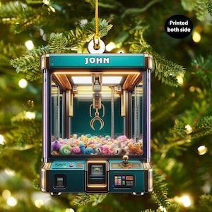 Puede incluir: Un adorno navideño de color verde azulado en forma de máquina de garras con detalles dorados. La máquina está llena de peluches de colores y tiene el nombre "JOHN" impreso en la parte delantera.