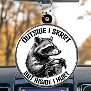 May include: A round, white car charm with a black and white illustration of a raccoon driving a car. The text "OUTSIDE I SKRRT BUT INSIDE I HURT" is printed in black around the raccoon.