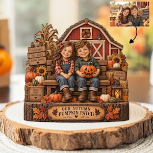 Puede incluir: Una figura decorativa de mesa con un fondo de granero rojo y dos niños sentados en un banco sosteniendo una calabaza. La base tiene la inscripción "OUR AUTUMN PUMPKIN PATCH" y está rodeada de calabazas, tallos de maíz y hojas de otoño.