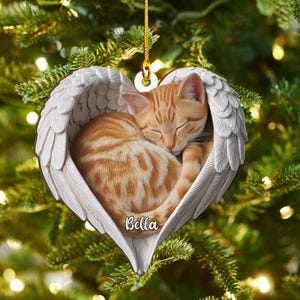 Op de afbeelding: Een hartvormige kerstversiering met een slapende oranje tabbykat genesteld in witte engelen vleugels. De naam van de kat, "Bella", staat op het ornament. Opgehangen aan een gouden draad.
