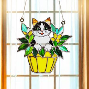 May include: A stained glass window hanging features a black and white cat sitting in a yellow flower pot with green leaves.