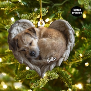 May include: A heart-shaped Christmas ornament featuring a sleeping dog nestled within angel wings. The ornament is tan and gray, with the name "Bella" printed on it. The text "Printed both side" is also visible.