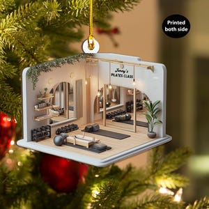 Könnte beinhalten: Ein Weihnachtsornament in Form eines Pilates-Studios mit Gewichten, Matten und Spiegeln. Das Ornament trägt den Text "Jenny's Pilates Class" und ist beidseitig bedruckt. Es hängt an einem goldenen Faden und vor einem Weihnachtsbaum.