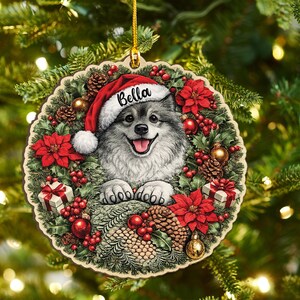 May include: A round wooden Christmas ornament featuring a fluffy grey dog wearing a red Santa hat with the name Bella printed on the brim. The dog is surrounded by a festive wreath of green pine needles, red poinsettias, pinecones, and gifts.