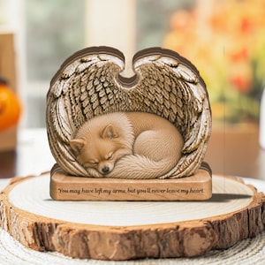 Pode incluir: Uma estatueta decorativa apresentando um cão castanho-claro enrolado a dormir, emoldurado por duas grandes asas de anjo detalhadas. A base está gravada com o texto "You may have left my arms, but you'll never leave my heart." e o nome "Bella".