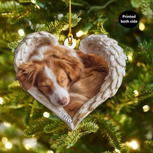 May include: Heart-shaped Christmas ornament featuring a sleeping brown and white dog nestled in angel wings. The ornament has the name "Bella" printed on it. The ornament is hanging from a gold string.