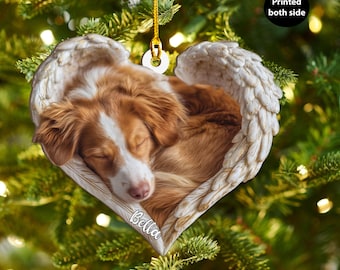 Può includere: Ornamento natalizio a forma di cuore con un cane marrone e bianco addormentato adagiato in ali d'angelo. L'ornamento ha il nome "Bella" stampato sopra. È appeso a un filo d'oro.