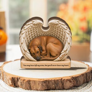May include: A decorative memorial ornament featuring a brown dog curled up inside a pair of large, detailed angel wings. The base is light wood with the text, "You may have left my arms, but you'll never leave my heart." and the name "Bella."