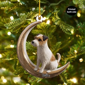 May include: A Christmas ornament featuring a Jack Russell Terrier dog sitting on a crescent moon. The moon is gold and white, with the dog having brown, black, and white fur. The word "Bella" is written on the moon. The ornament hangs from a gold string.