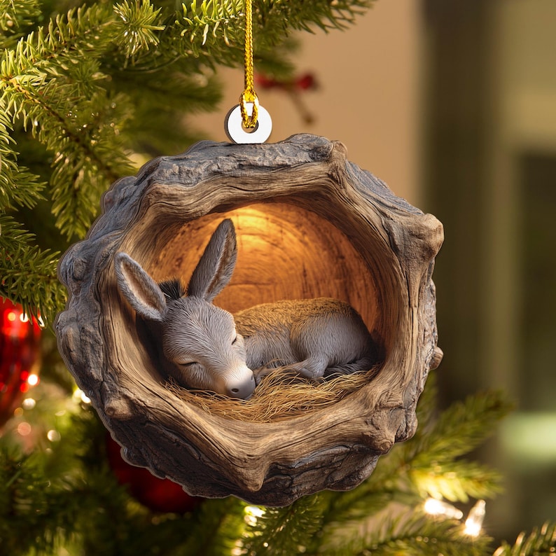May include: A Christmas ornament featuring a sleeping donkey nestled inside a wooden hollow. The donkey is gray with closed eyes, resting on a bed of straw. The ornament is suspended by a gold string, with a white circular accent.