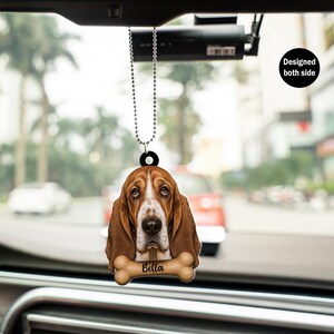 May include: A car hanging ornament featuring a Basset Hound dog with brown and white fur. The dog's name, "Bella", is printed on a bone-shaped design. The ornament is designed on both sides and hangs from a silver beaded chain.