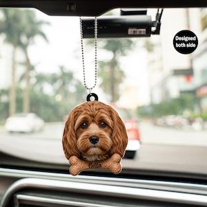 Op de afbeelding: Een auto-hangornament met een gedetailleerde afbeelding van het gezicht van een bruine Cavapoo-hond. De naam van de hond, "Bella", is gedrukt op een botvormig accent. Het ornament is aan beide zijden ontworpen en hangt aan een kralenketting.
