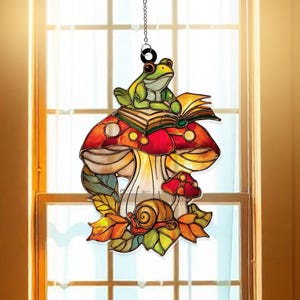 May include: Stained glass window hanging featuring a green frog atop a red and orange mushroom with an open book. A snail rests on leaves below. The whimsical design is suspended by a chain, perfect for a window or wall.
