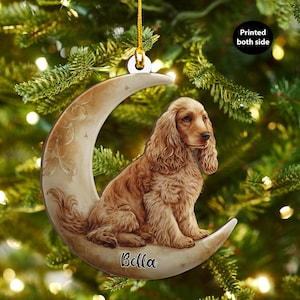 Op de afbeelding: Een kerstversiering met een Cocker Spaniel hond die op een maan zit. De versiering is lichtbruin, met de naam van de hond, "Bella", op de maan gedrukt. Het hangt aan een gouden touwtje tegen een achtergrond van groene kerstboomtakken.