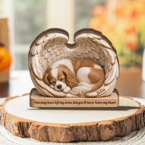 Puede incluir: Una figura decorativa que muestra a un cachorro de spaniel marrón y blanco durmiendo, acurrucado entre dos grandes alas de ángel texturizadas. La base tiene grabado el texto: "You may have left my arms, but you'll never leave my heart."