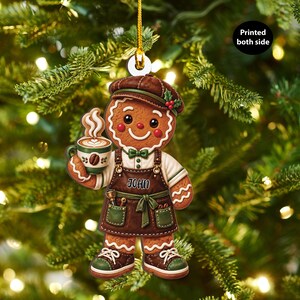 May include: A gingerbread person ornament wearing a brown cap, white shirt, and brown apron with the name JOHN printed on it. The figure holds a green coffee mug with steam rising. The ornament features a gold hanging string and text Printed both side.