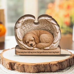 May include: A tan puppy figurine curled up inside a pair of large, detailed angel wings. The base features the engraved text, "You may have left my arms, but you'll never leave my heart," and the name "Bella" is inscribed on the wing.
