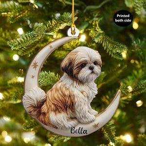 Può includere: Un ornamento natalizio raffigurante un cane Shih Tzu seduto su una luna crescente. Il cane ha il pelo marrone e bianco, e la luna è color crema con accenti di stelle. Il nome "Bella" è scritto sulla luna. L'ornamento è appeso a un filo d'oro.