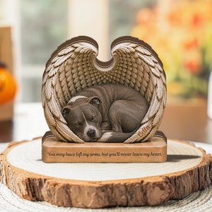 Puede incluir: Un adorno conmemorativo decorativo que muestra a un perro pitbull atigrado durmiendo acunado por un par de alas de ángel grandes y detalladas. La base tiene grabada la frase "You may have left my arms, but you'll never leave my heart" y el nombre "Bella".