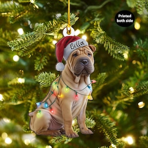May include: A Christmas ornament featuring a Shar Pei dog wearing a Santa hat with the name "Bella" and a string of colorful Christmas lights. The ornament is printed on both sides and hangs from a gold string.