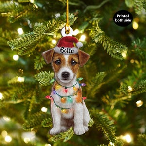 May include: A Christmas tree ornament featuring a Jack Russell Terrier wearing a Santa hat with the name "Bella" and a string of colorful Christmas lights. The ornament is printed on both sides and hangs from a gold cord.