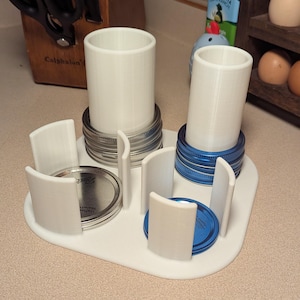 May include: A white 3D-printed jar lid and jar holder. The holder has two cylindrical sections for jars, surrounded by curved dividers. The holder also has spaces for jar lids. The lids are silver and blue.